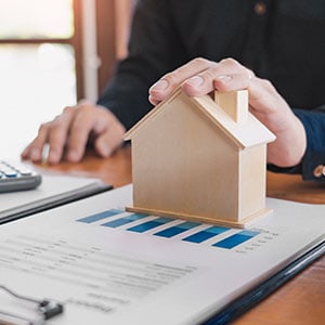 Hands holding wooden house model above property agreement with bar graphs; financial planning scene on a wooden desk in natural light.